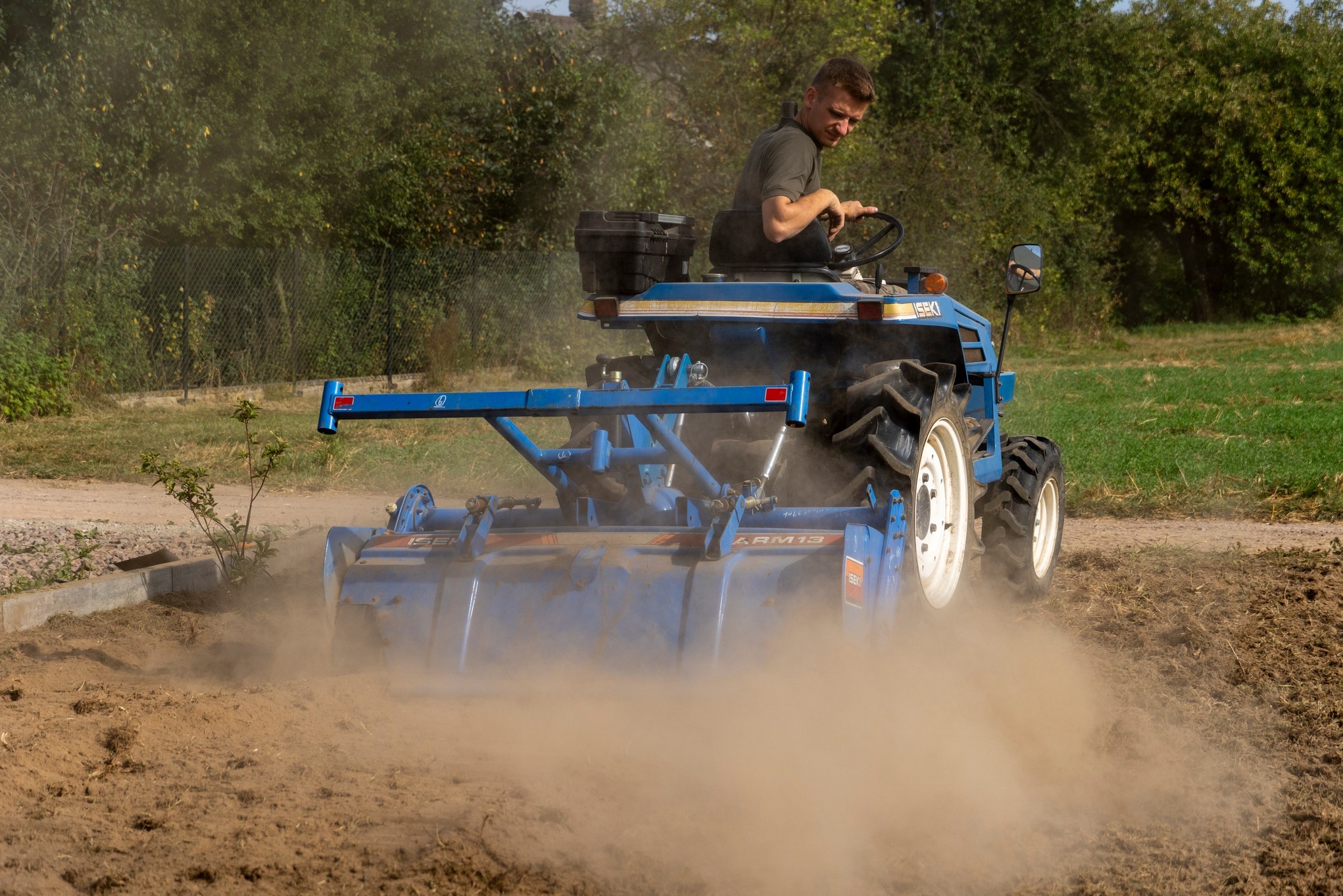 Niebieski traktor ISEKI z glebogryzarką spulchnia ziemię na polu, za nim zielone drzewa. Operator skupiony na pracy. Przygotowanie terenu pod uprawę.