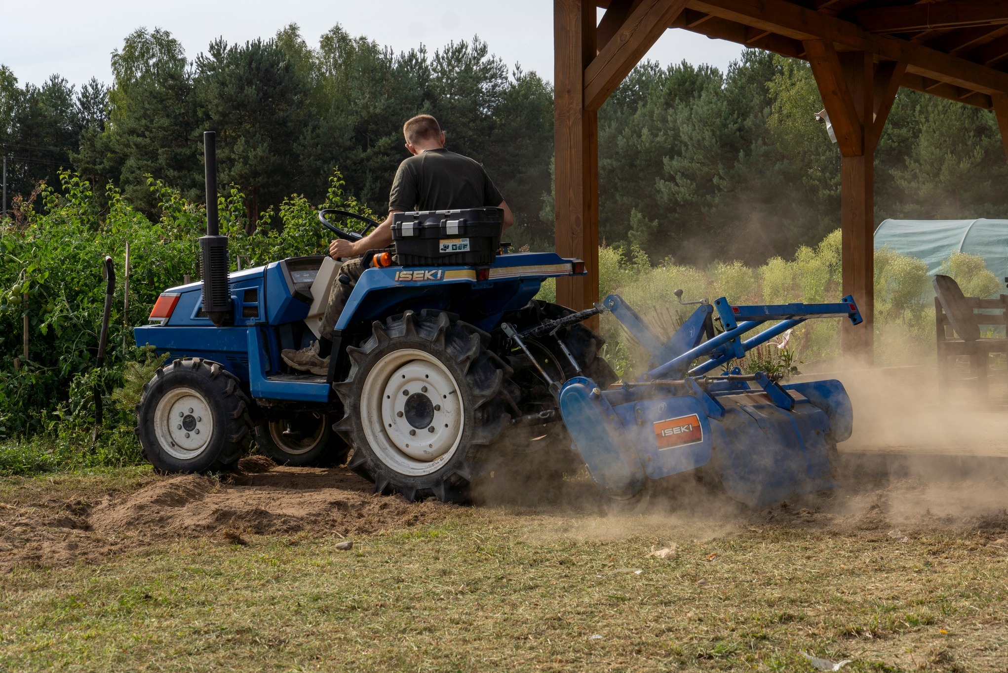 Niebieski traktor Iseki w trakcie pracy na polu, z widocznym pyłem i świeżo przekopaną ziemią. Operator siedzi na maszynie, w tle las i drewniana konstrukcja.