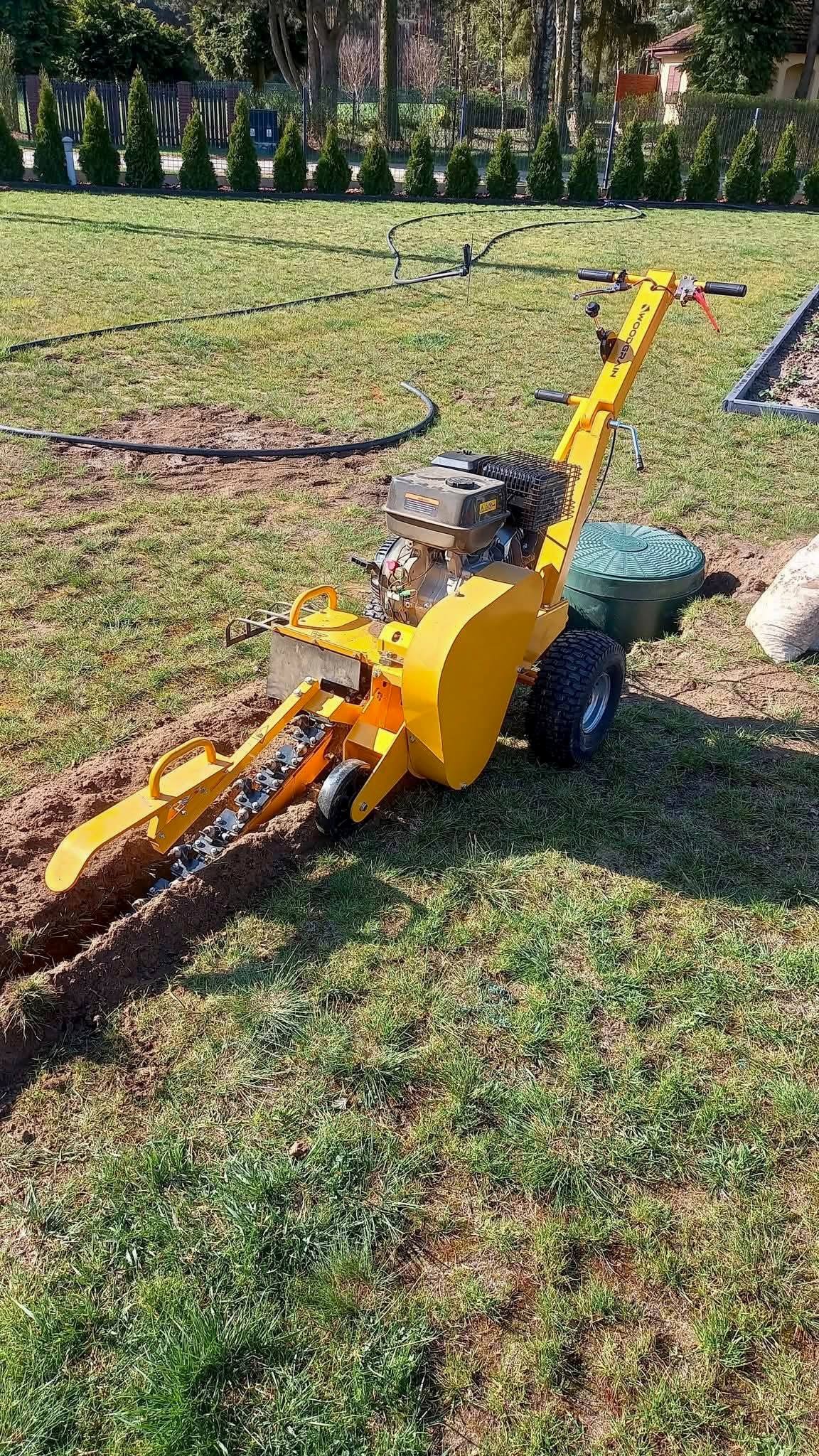 Żółta glebogryzarka w trakcie pracy w Szydłowcu, tworząca wąski wykop w trawniku. W tle widoczne tuje i czarny wąż nawadniający.