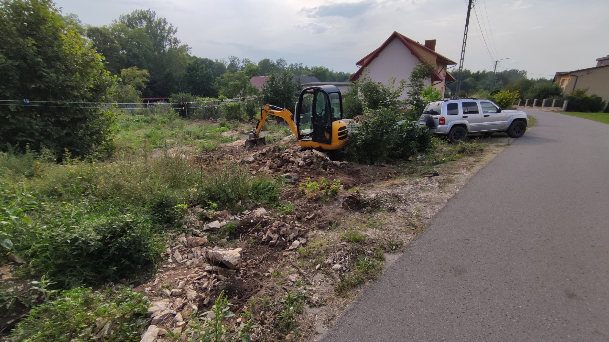 Żółta mini koparka na placu budowy, przygotowująca teren pod instalacje, z widocznymi kamieniami i wykopaną ziemią, w tle dom jednorodzinny i samochód osobowy.