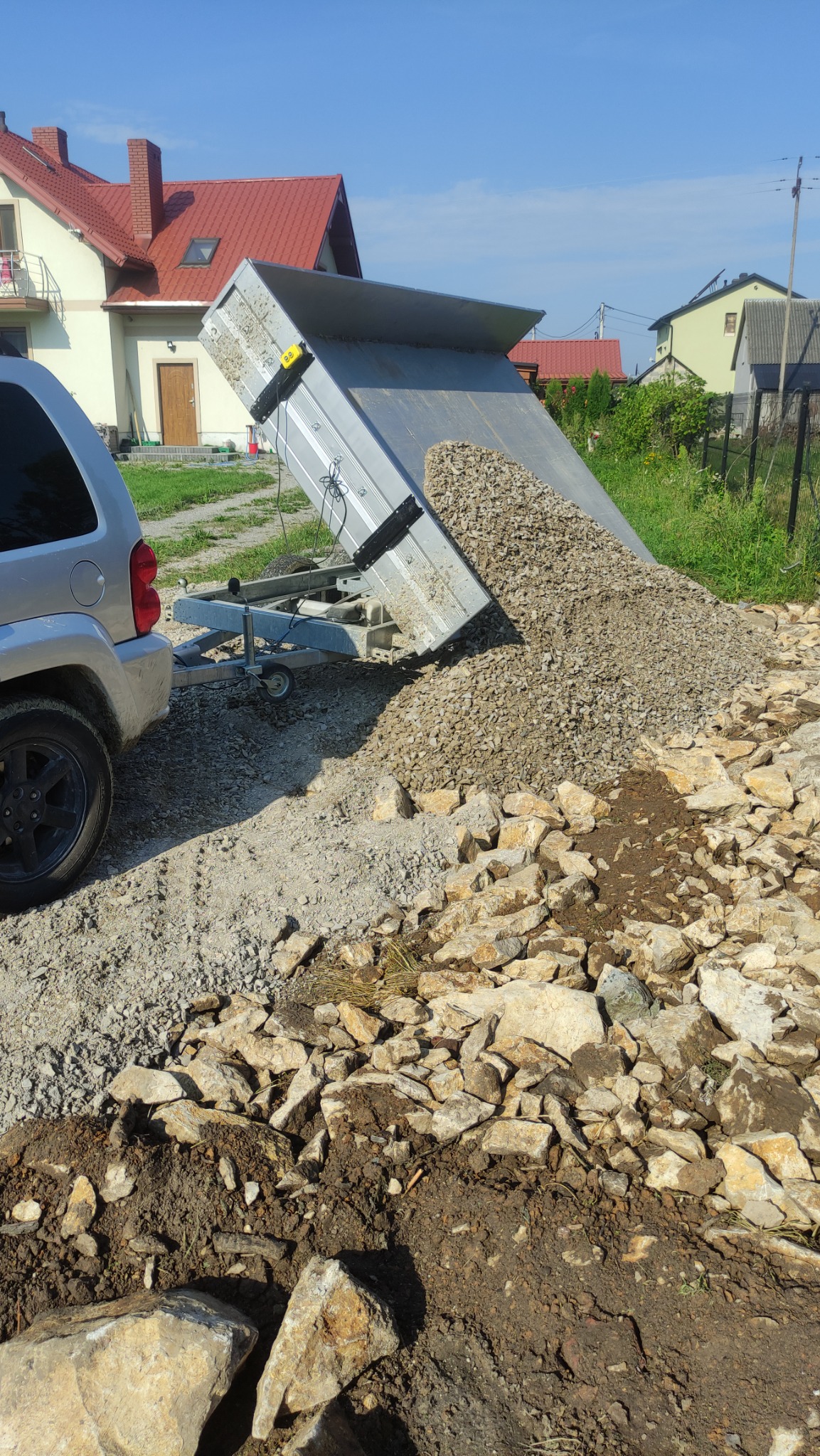 Przyczepa wywrotka z wysypanym żwirem na przygotowanym terenie pod budowę, widoczny samochód osobowy i zabudowania w tle.