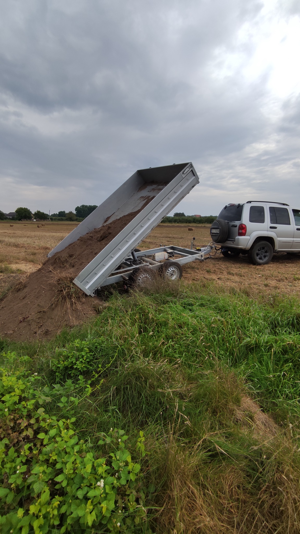 Przyczepa wywrotka z ziemią, podłączona do srebrnego samochodu terenowego na polu ze ścierniskami, zarośniętym trawą i chmurami na niebie.