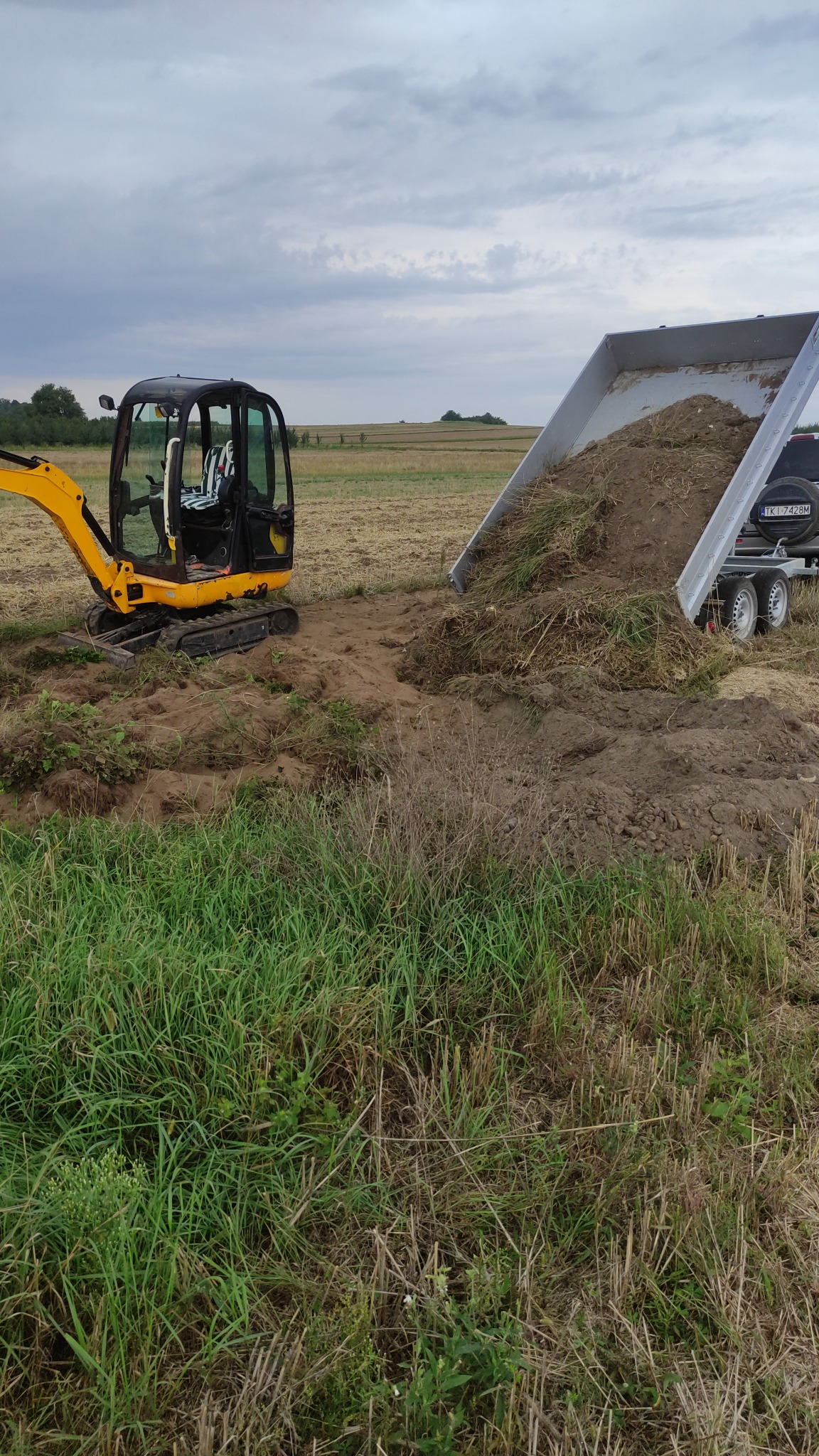 Żółta mini koparka obok przyczepy z wywrotką, która wysypuje ziemię i resztki roślin na pole z niską trawą, widoczne niebo z chmurami.
