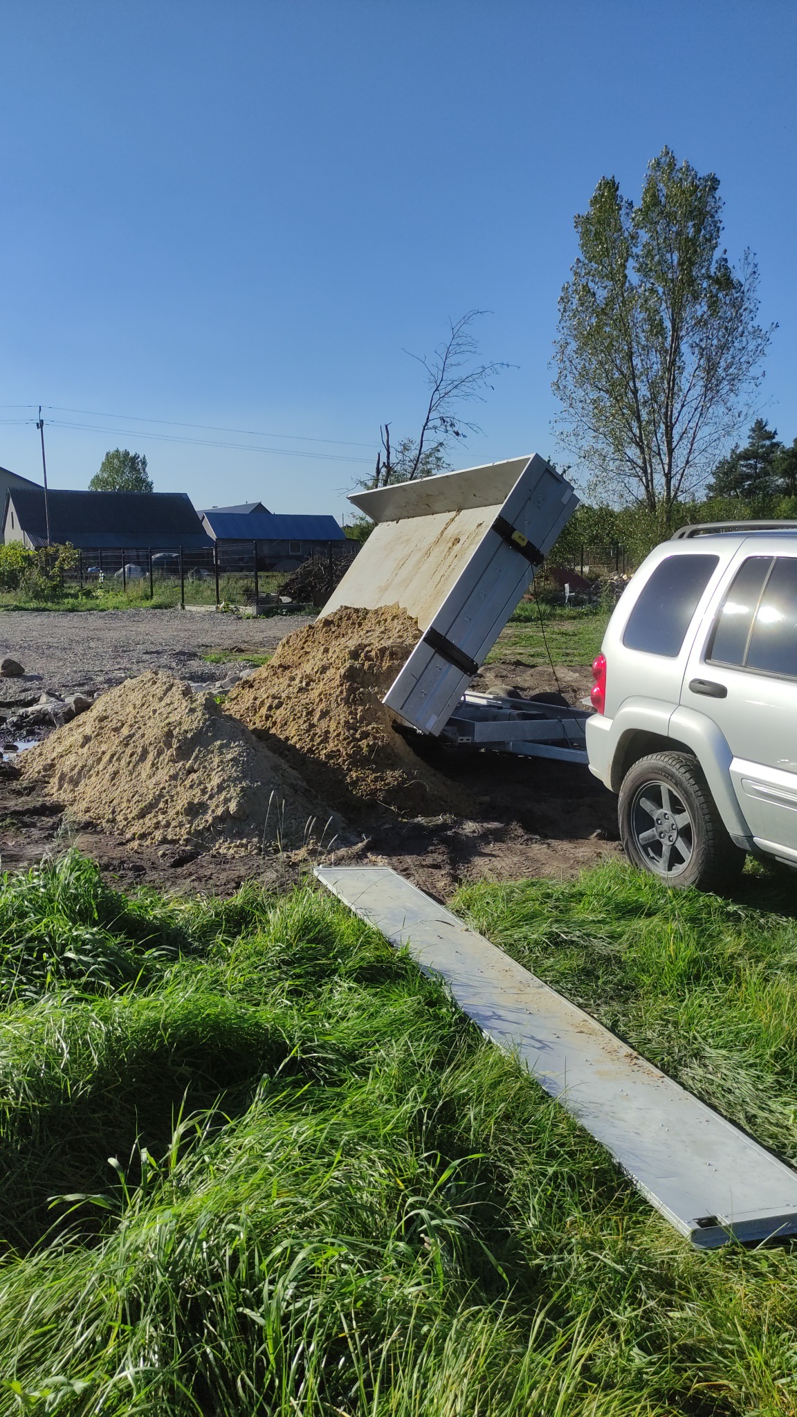 Przyczepa wywrotka z piaskiem na trawiastym terenie budowy, widoczny samochód terenowy i zabudowania w tle.