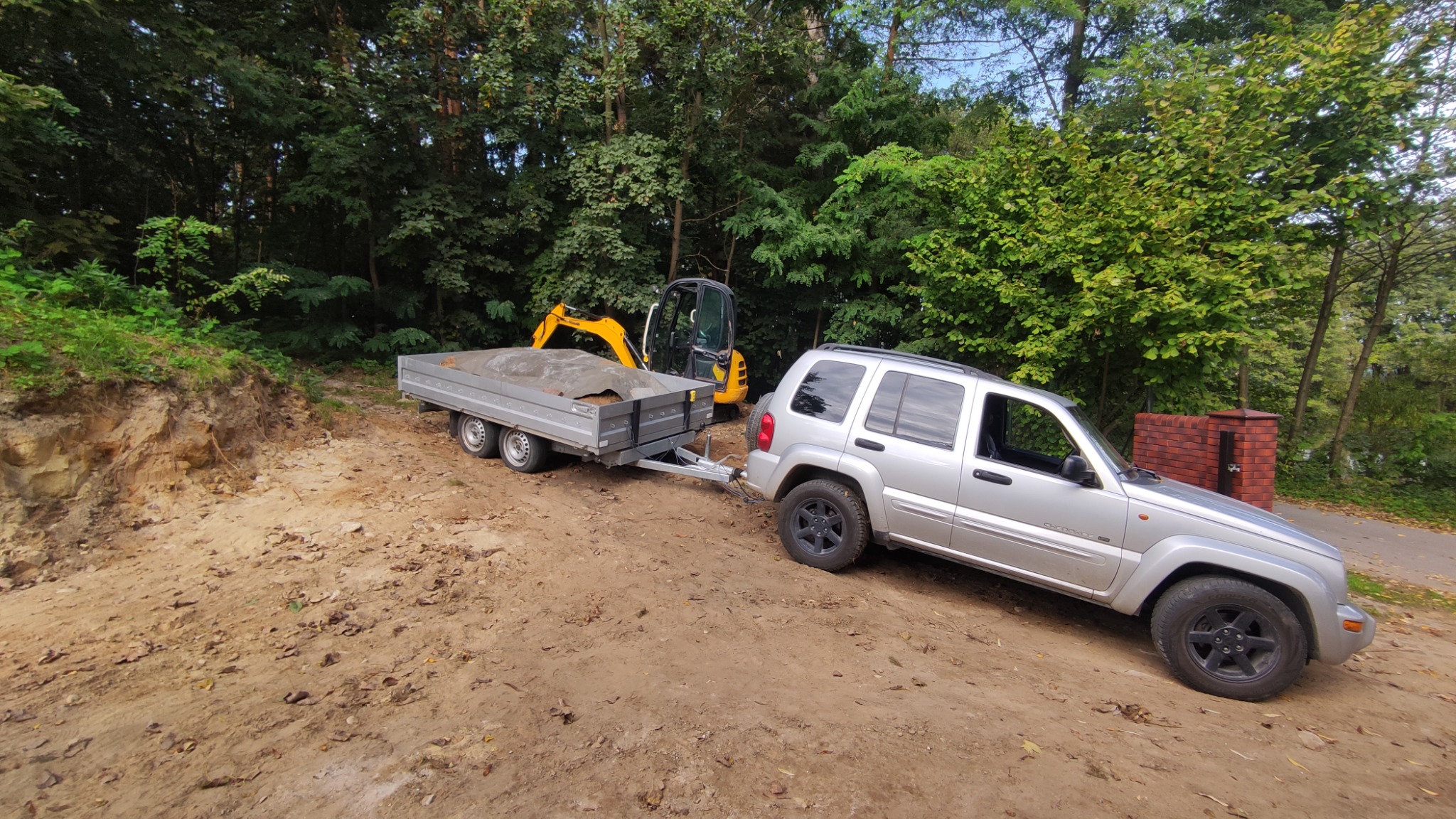 Srebrny samochód terenowy z przyczepą załadowaną ziemią, obok zaparkowana mała koparka na tle zieleni.