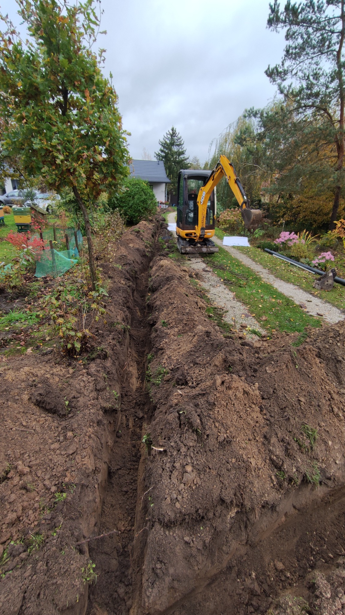 Wąski wykop w ziemi, wykonany minikoparką JCB na działce z drzewami i krzewami, przygotowany pod instalacje, widoczne rury i biała folia.