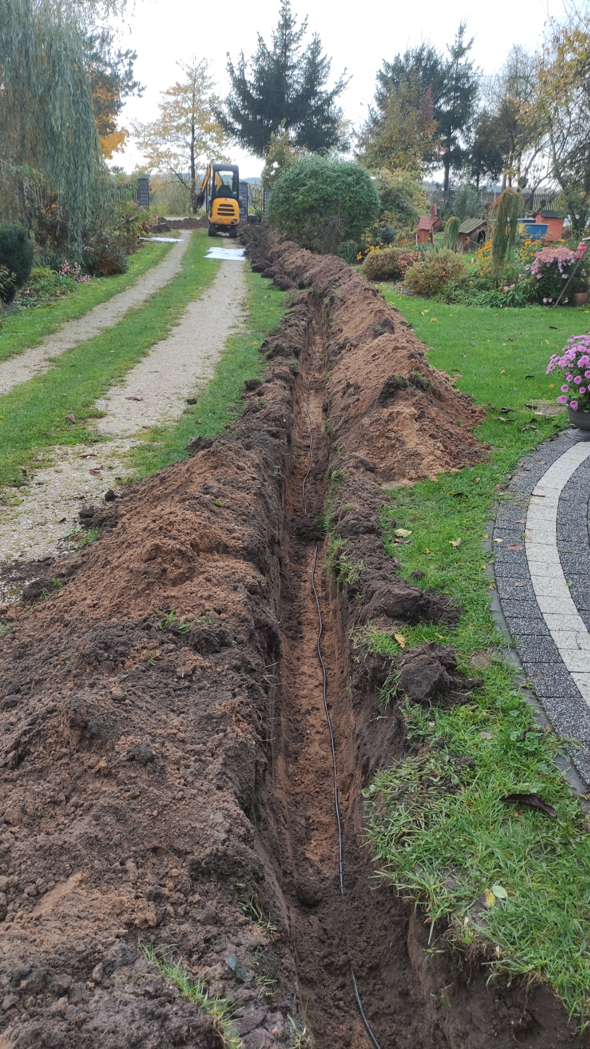 Wykopany wąski rów w ziemi wzdłuż trawnika i ścieżki, z widocznym kablem na dnie, w tle żółta koparka i drzewa.