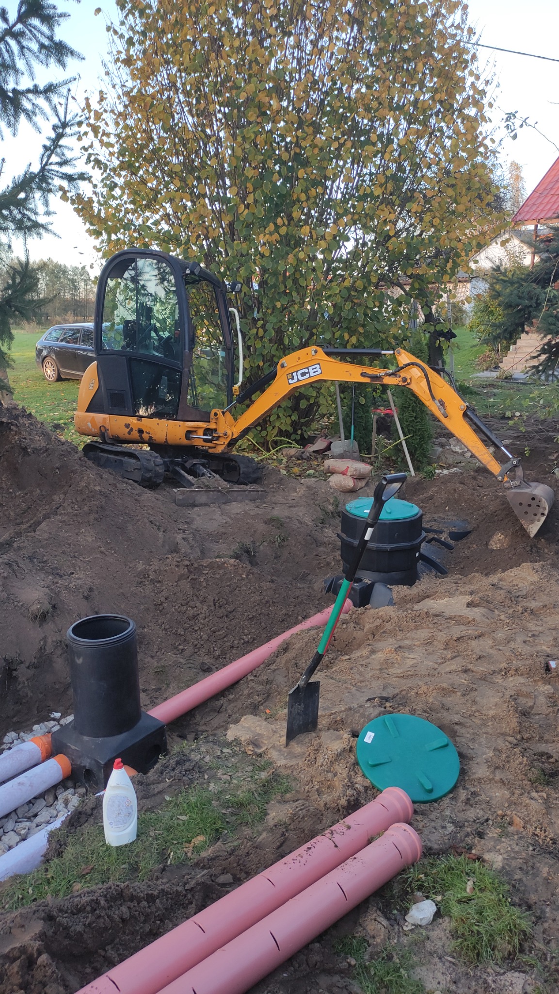 Żółta mini koparka JCB przy wykopie z rurami kanalizacyjnymi i studzienką, widoczna łopata oraz butelka płynu do naczyń, w tle drzewo i dom.