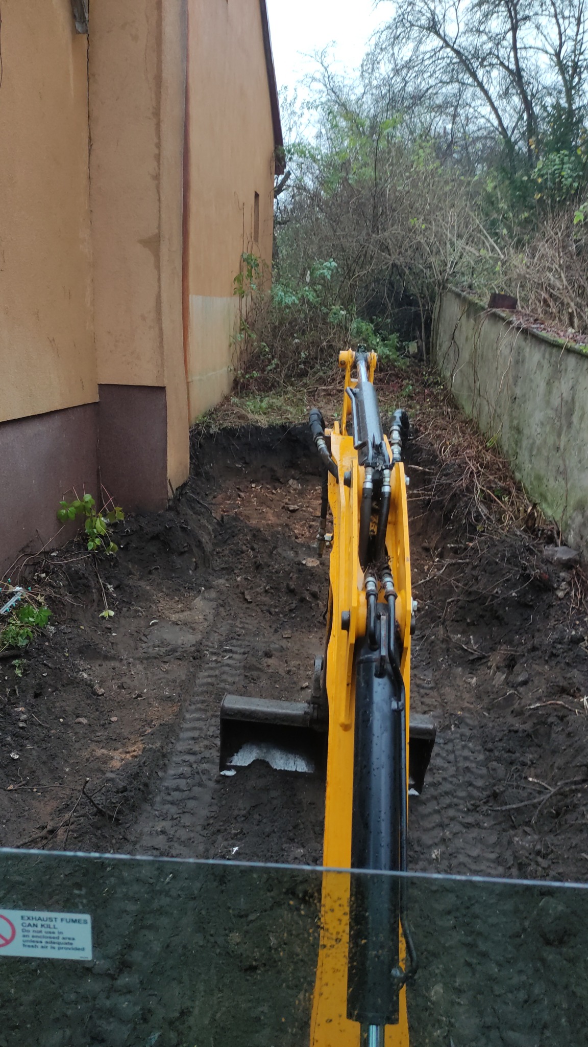 Żółto-czarna koparka wykonująca wykop w wąskiej przestrzeni między budynkiem a murem oporowym, widok z kabiny operatora, mokra ziemia i ślady opon.