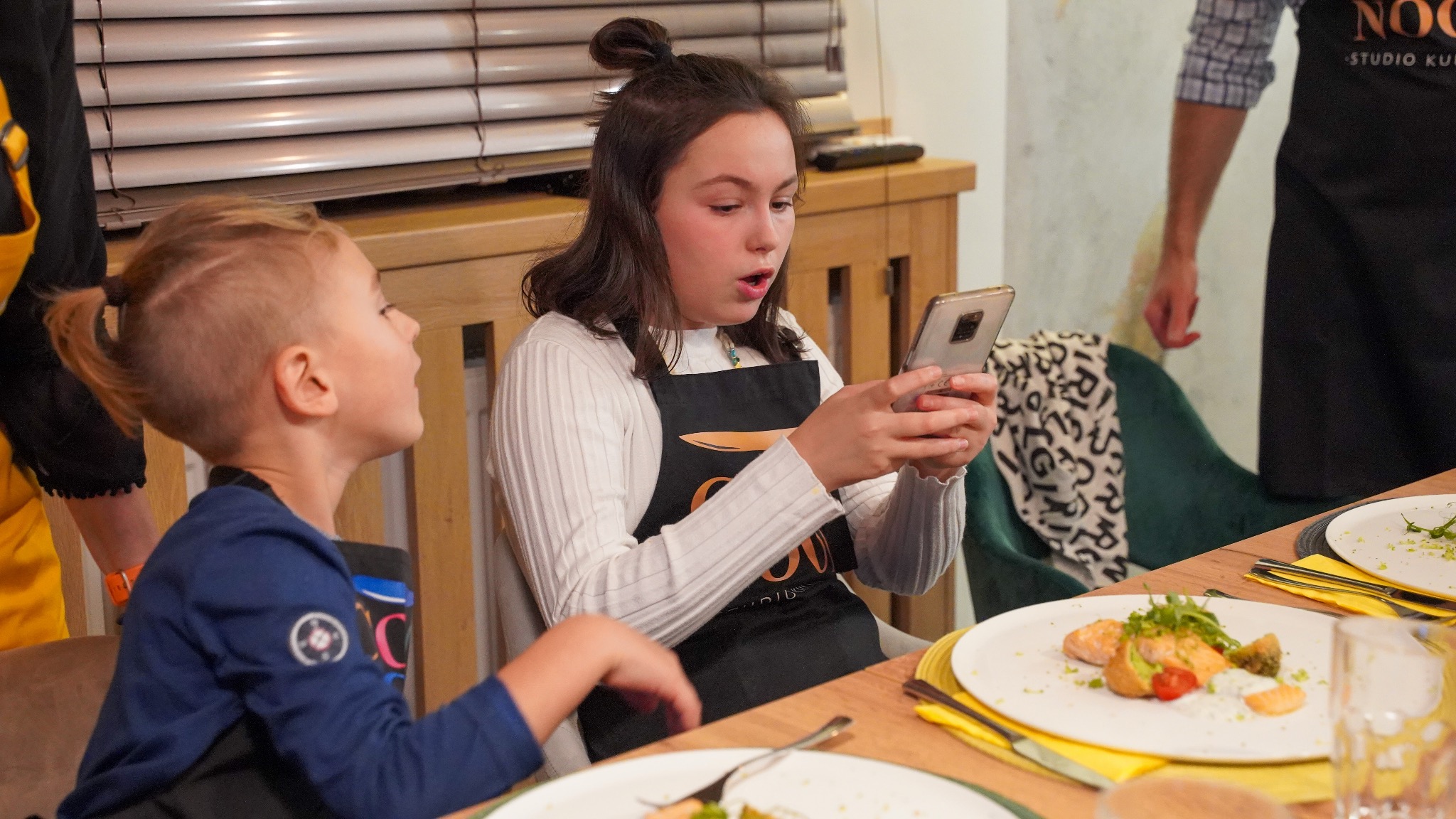 Dziewczynka w czarnym fartuchu z logo studia kulinarnego, z zaskoczeniem patrzy na telefon, obok niej mały chłopiec i talerz z daniem z łososia, w tle widać fragment wnętrza studia kulinarnego.