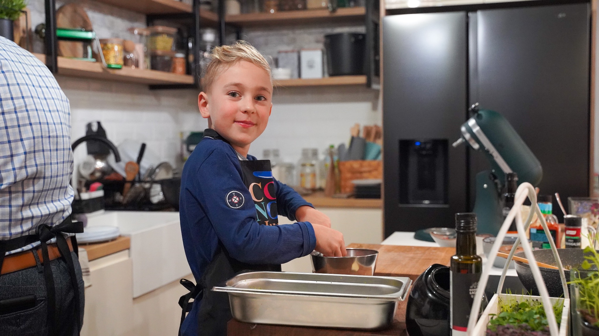 Uśmiechnięty chłopiec w fartuchu miesza składniki w metalowej misce w nowoczesnej kuchni, obok dorosły w koszuli w kratę, w tle czarna lodówka i mikser.
