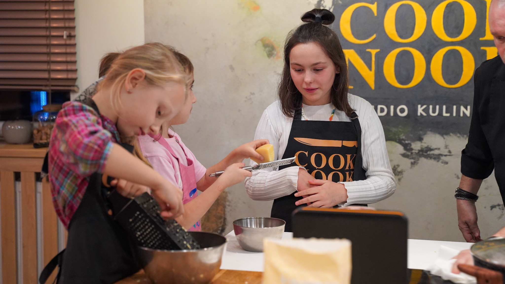 Dzieci i instruktor w fartuchach w studiu kulinarnym, dziewczynka trzyma ser i tarkę, druga ściera ser do miski, w tle logo studia kulinarnego.