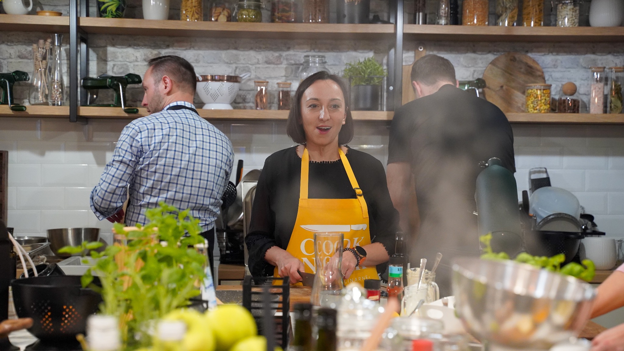 Kobieta w żółtym fartuchu kroi na desce w kuchni podczas warsztatów kulinarnych, w tle półki z słoikami i sprzętem kuchennym oraz inne osoby.
