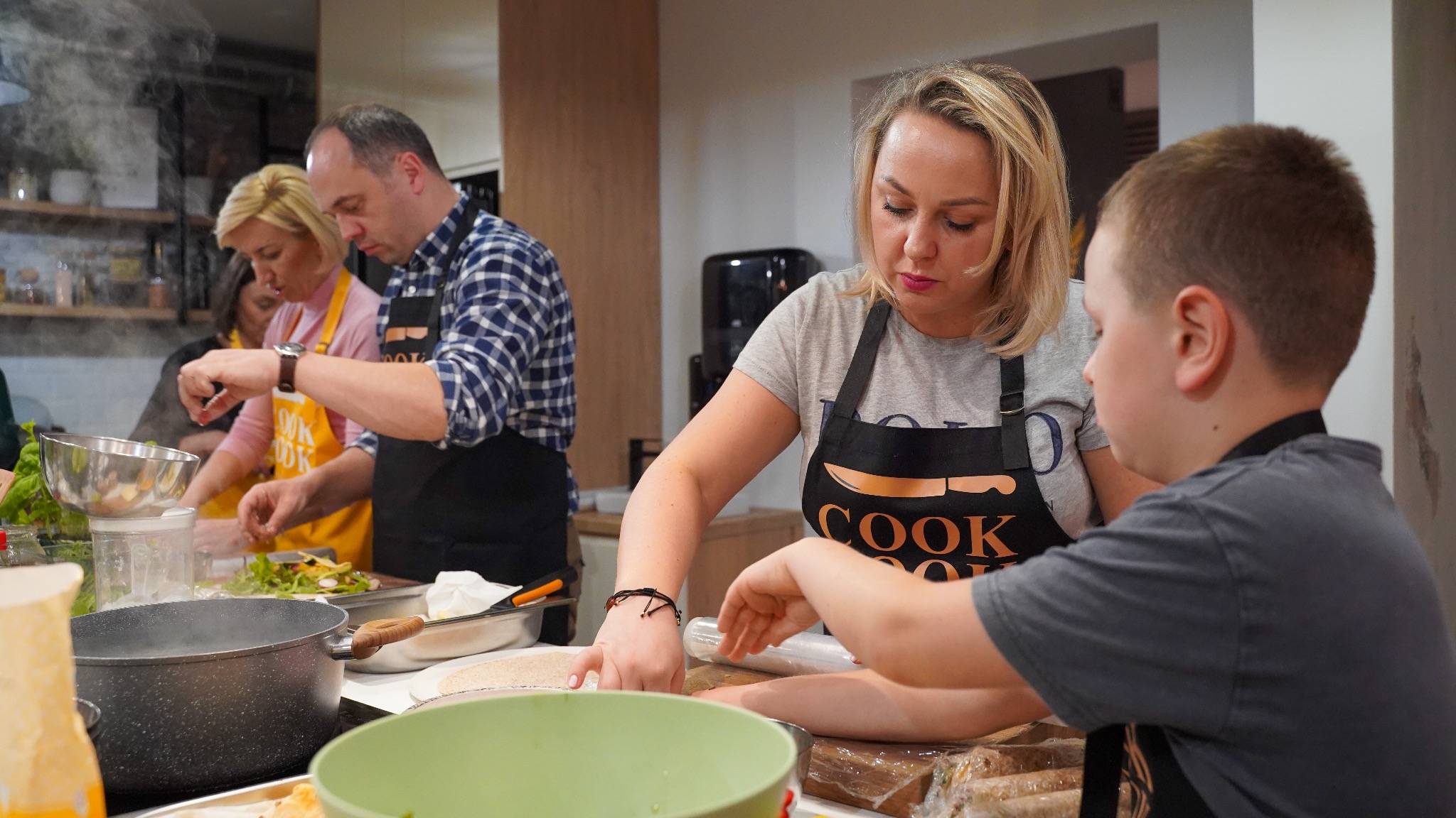Grupa osób, w tym kobieta z blond włosami w czarnym fartuchu z napisem 'COOK', przygotowuje jedzenie w kuchni, inni uczestnicy w tle kroją warzywa i mieszają składniki.