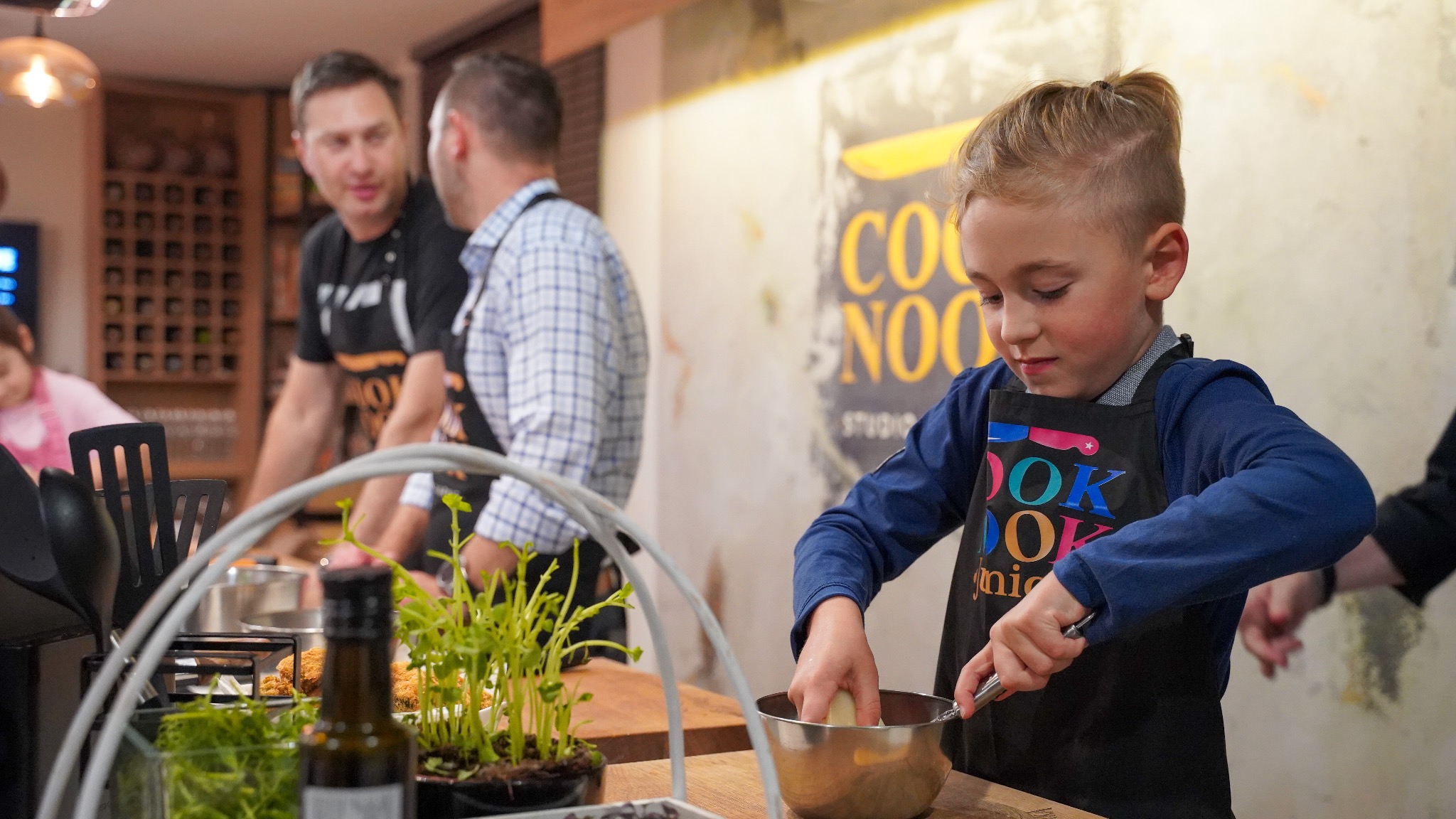 Chłopiec w czarnym fartuchu z kolorowym napisem 'COOK' miesza składniki w metalowej misce, w tle dwóch mężczyzn rozmawia, widoczny plakat 'COOK NOOK studio' i dekoracje roślinne na stole.