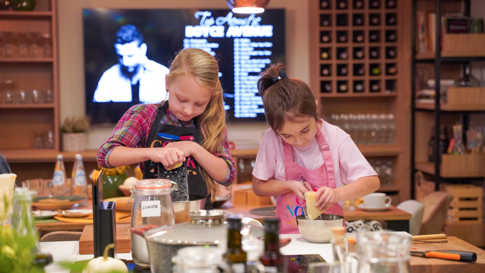 Dwie dziewczynki w fartuchach ścierają ser na tarce w kuchni podczas warsztatów kulinarnych; w tle widoczny ekran z nagraniem wideo.