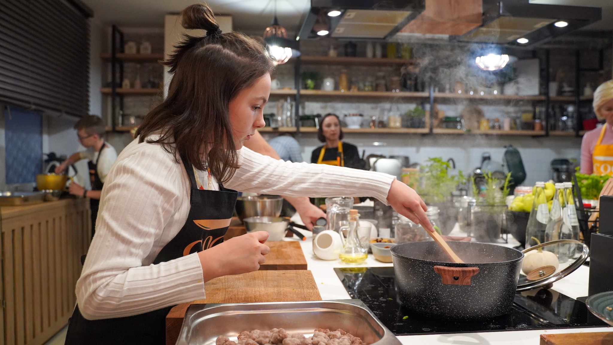 Młoda kobieta w fartuchu miesza drewnianą łyżką w garnku na płycie indukcyjnej podczas warsztatów kulinarnych, w tle inni uczestnicy i półki z produktami.
