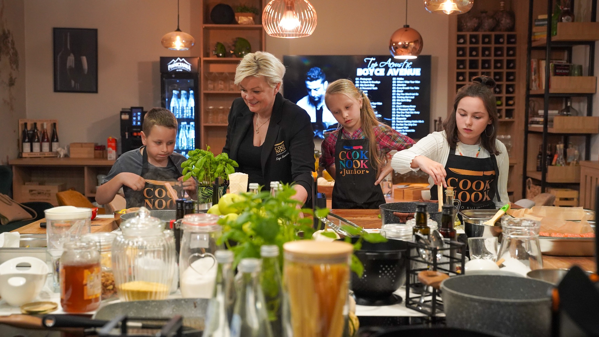 Warsztaty kulinarne dla dzieci i dorosłych w studio Cook Nook: kobieta prowadząca zajęcia i troje dzieci przy stole pełnym składników i naczyń, w tle monitor z listą utworów Top Acoustic Boyce Avenue.