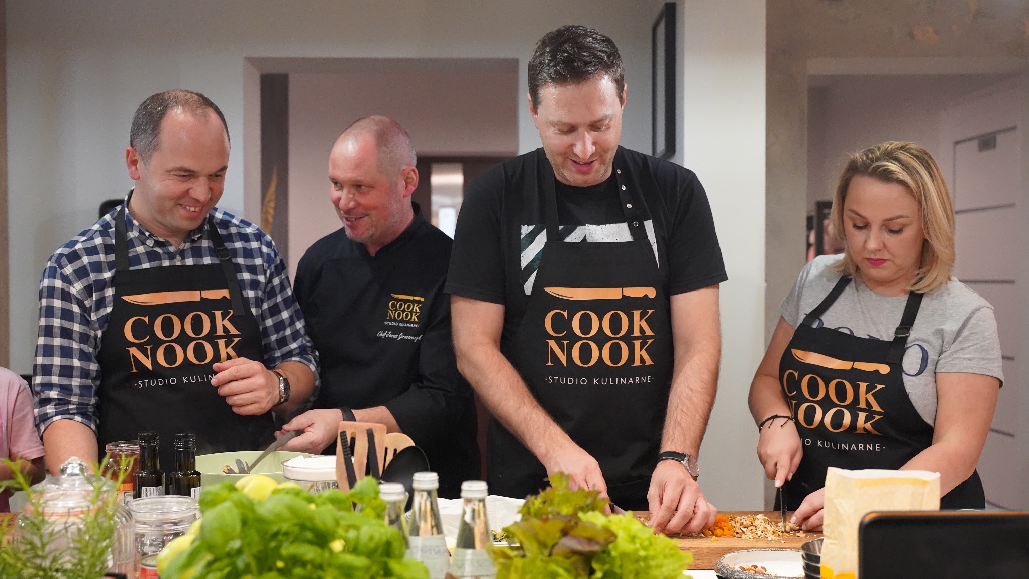 Cztery osoby w fartuchach z logo studia kulinarnego Cook Nook przygotowują posiłek, krojąc warzywa i orzechy na drewnianych deskach w jasnym wnętrzu kuchni.