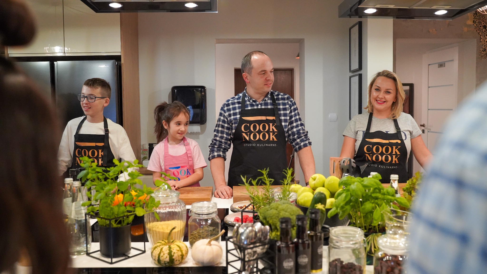 Grupa osób w fartuchach 'COOK NOOK Studio Kulinarne' stoi za stołem w kuchni, na którym znajdują się warzywa, owoce, przyprawy i dekoracje roślinne.