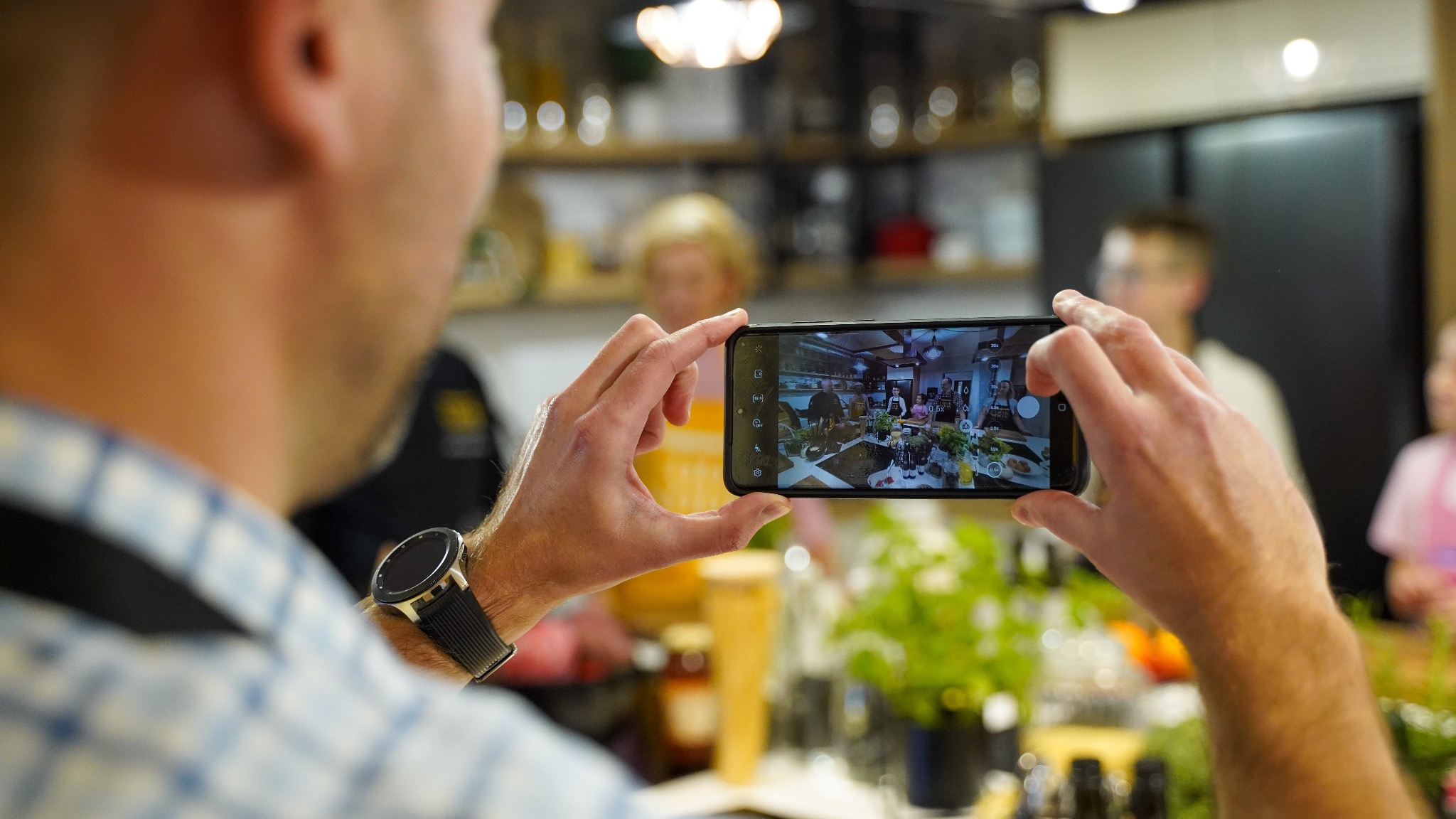 Osoba fotografuje telefonem grupę ludzi przy stole z jedzeniem i napojami w jasnym pomieszczeniu, widoczny zegarek na nadgarstku.