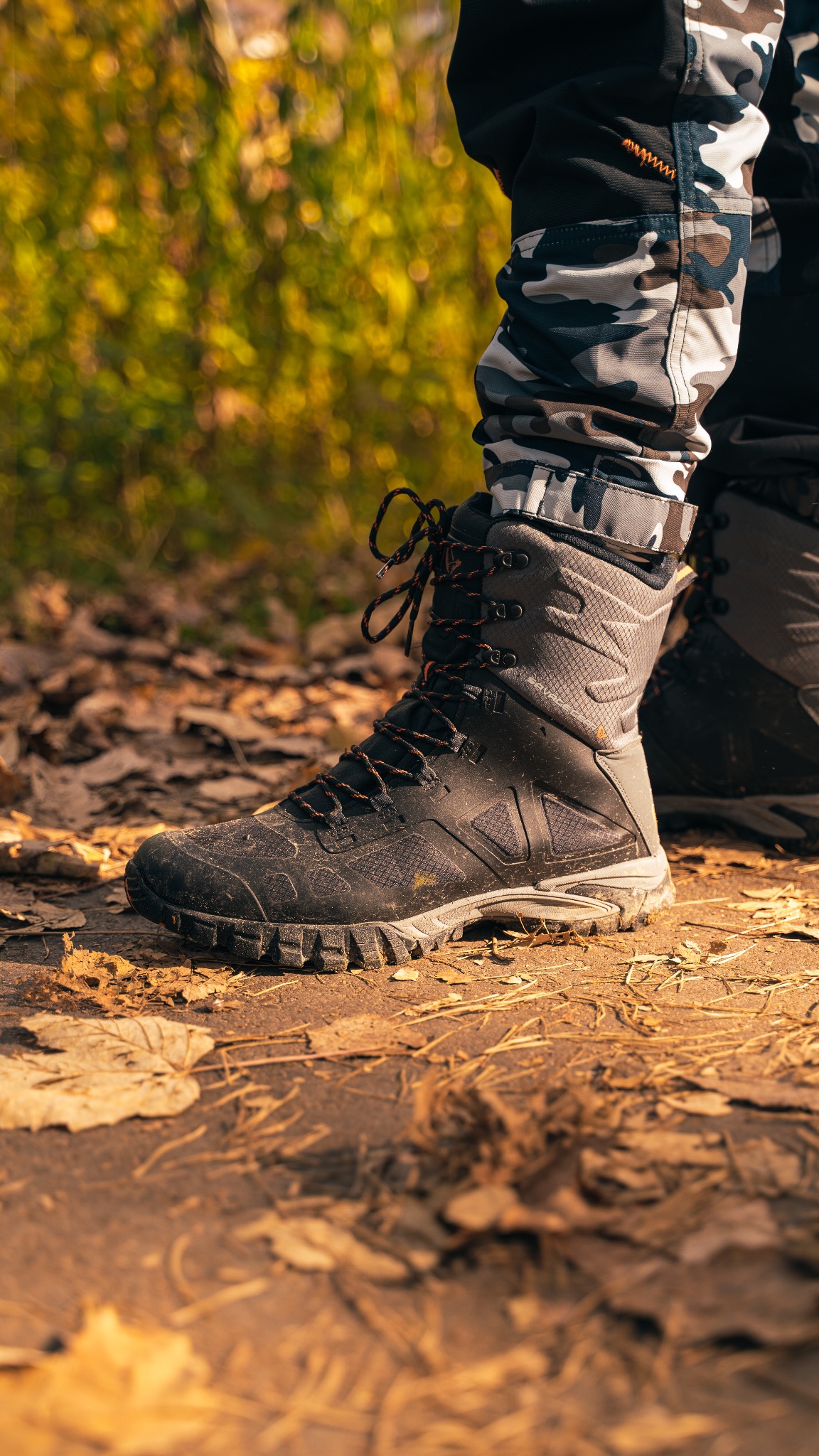 Czarny, sznurowany but trekkingowy z pomarańczowymi sznurówkami, noszony przez osobę w spodniach moro, stojącą na ziemi pokrytej liśćmi w jesiennym lesie.