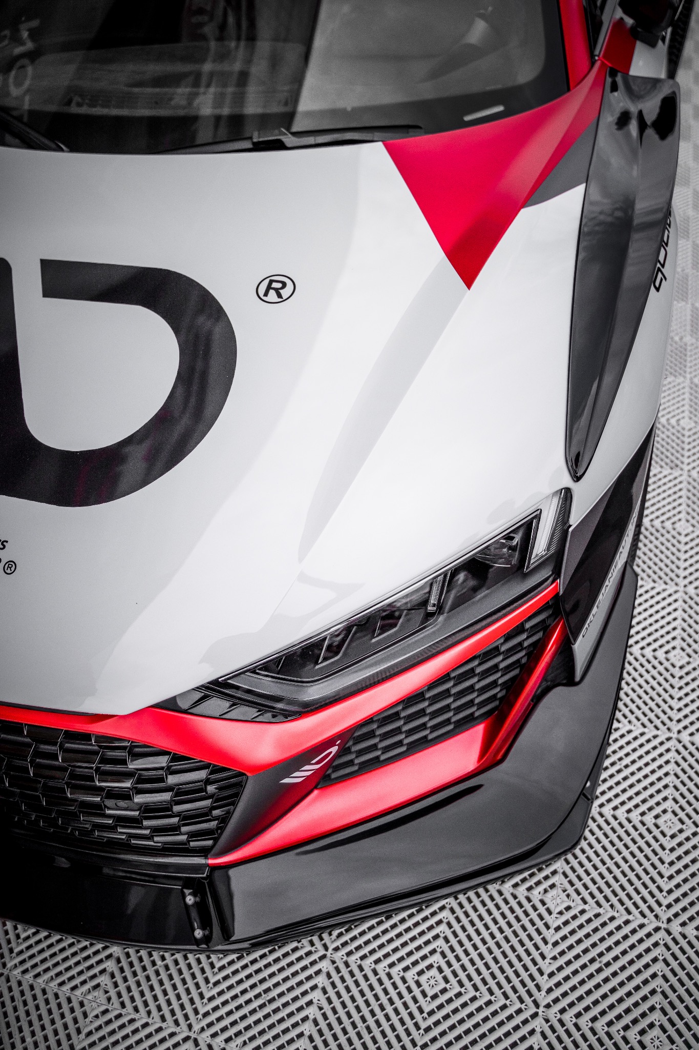 Fragment przedniej części sportowego samochodu z białym lakierem, czerwonymi akcentami i czarnymi elementami aerodynamicznymi, widoczne logo i numer na karoserii