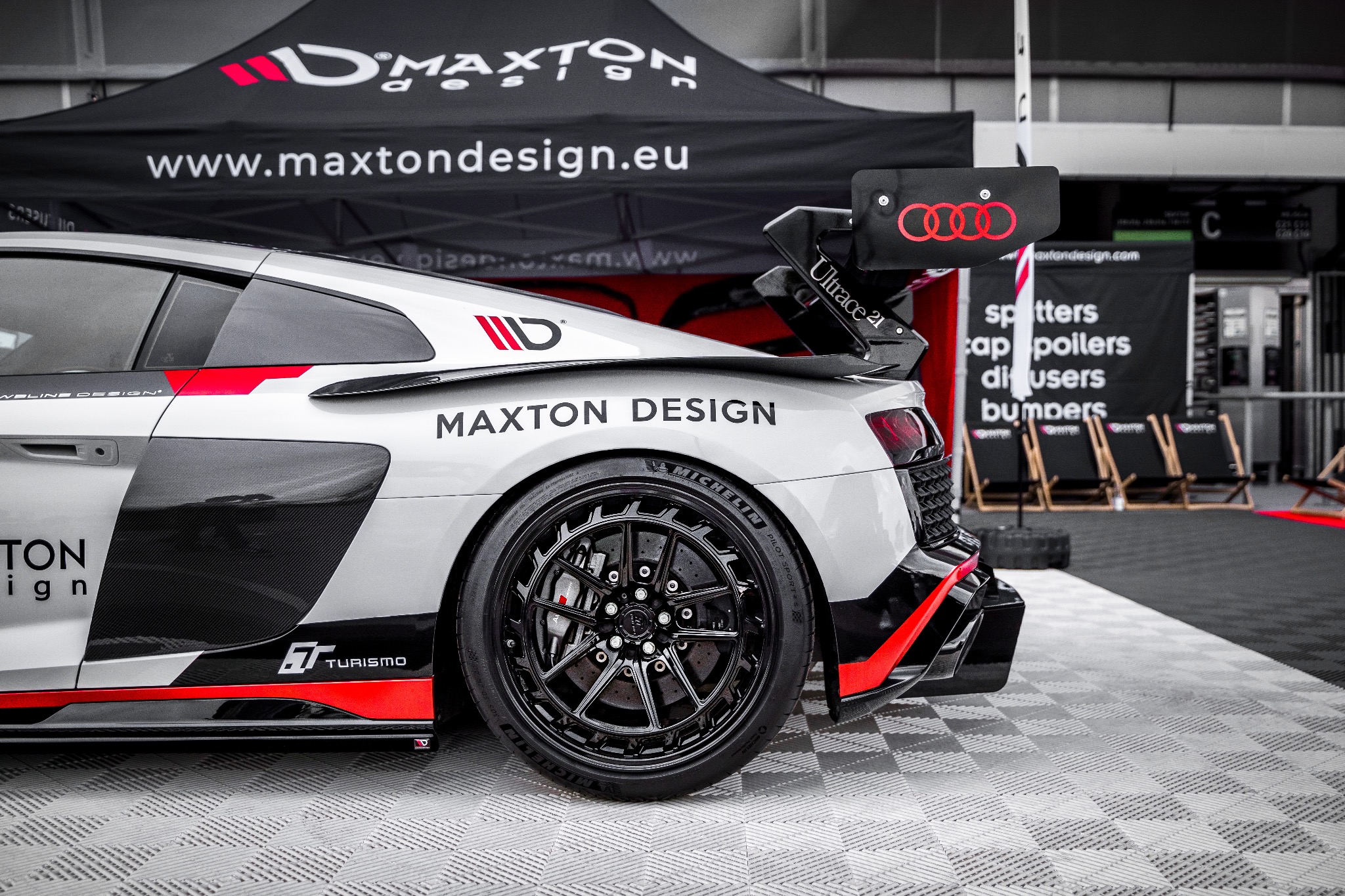 Szary sportowy samochód Audi R8 z czarnymi felgami i czerwonymi akcentami, zaparkowany na szarej podłodze w kratkę, z logotypami firm tuningowych Maxton Design i Ultrace 21, na tle namiotu...