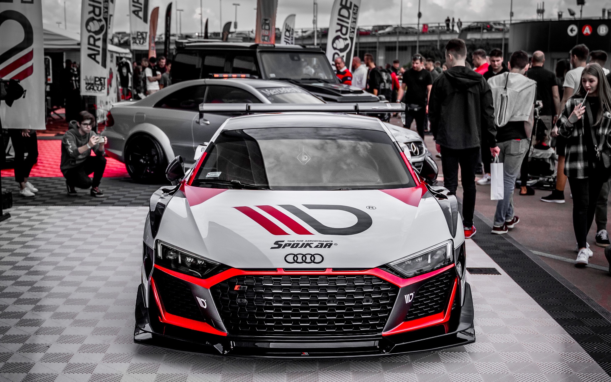 Białe Audi R8 z czerwonymi akcentami i logo 'SPOJKAR' na masce, prezentowane na tle tłumu ludzi i stoisk podczas motoryzacyjnej imprezy plenerowej.