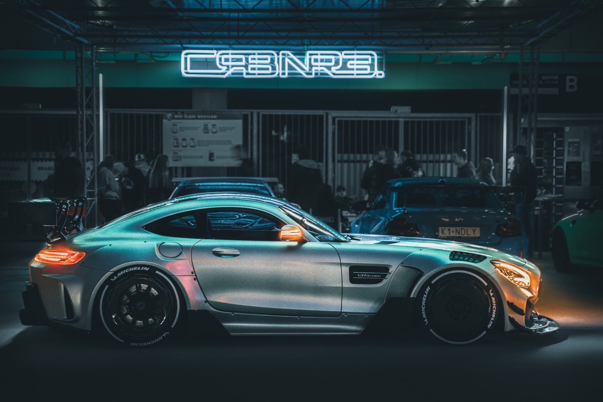 Srebrny sportowy samochód z tuningiem, szerokimi oponami z napisem Michelin, na tle neonu CRBNRE i innych pojazdów w hali wystawienniczej.