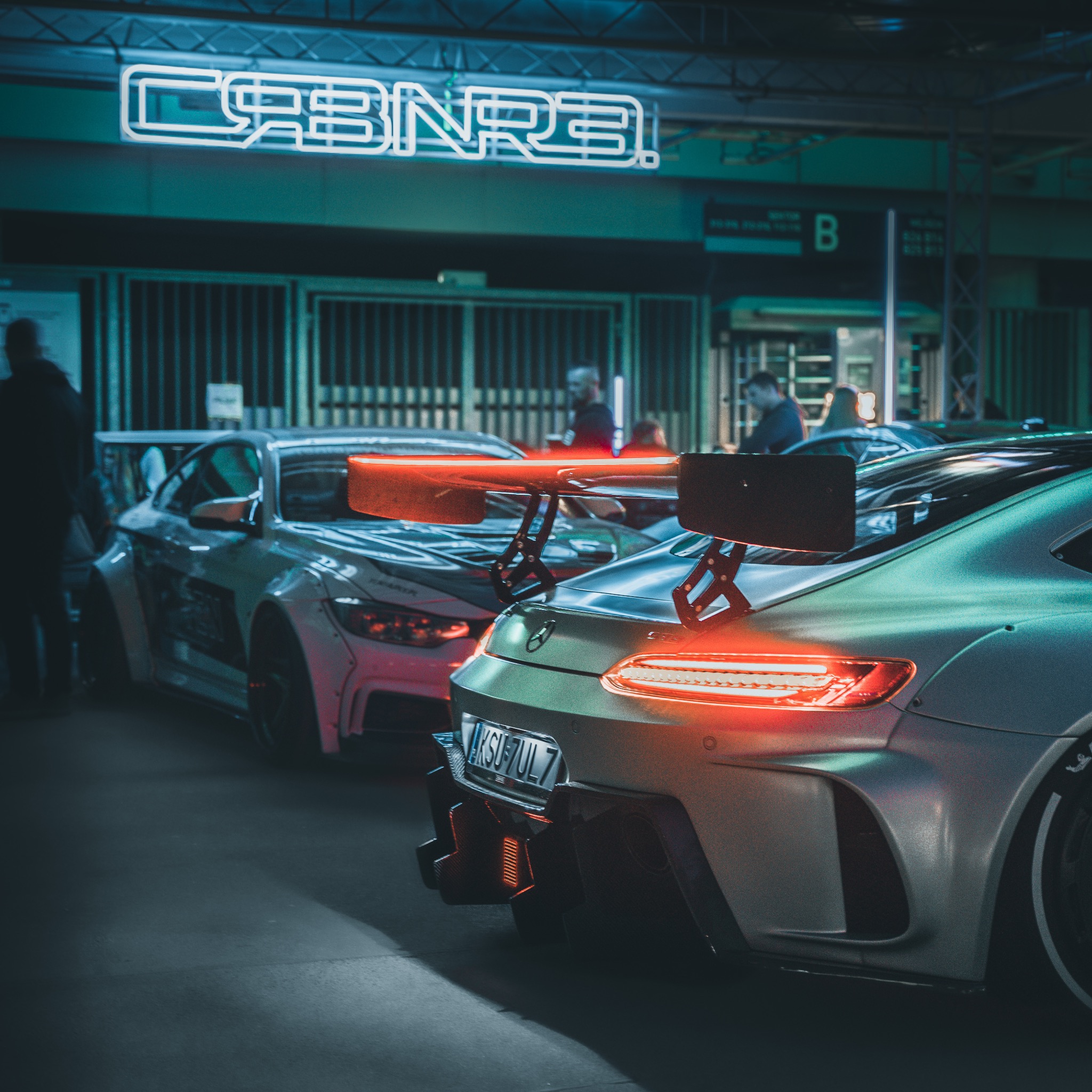 Dwa zmodyfikowane samochody sportowe z dużymi spojlerami na wystawie, oświetlone neonowym znakiem z tyłu.