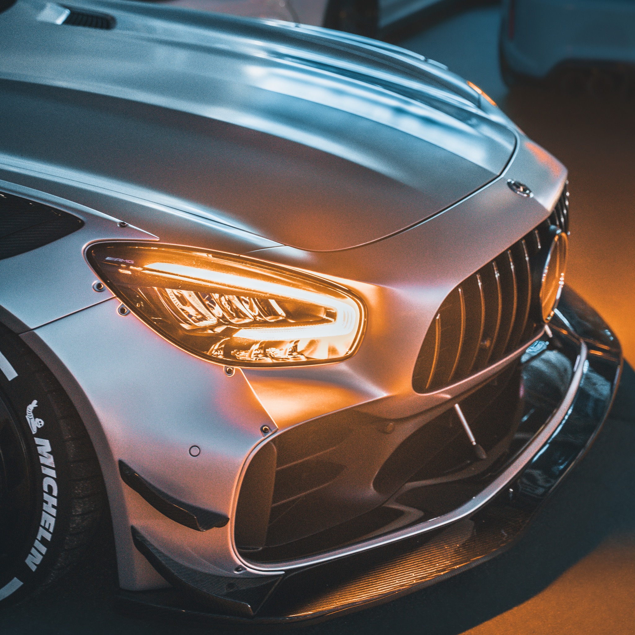 Przód srebrnego samochodu sportowego AMG z włączonymi światłami LED, widoczne detale karoserii i opona Michelin z białym napisem.