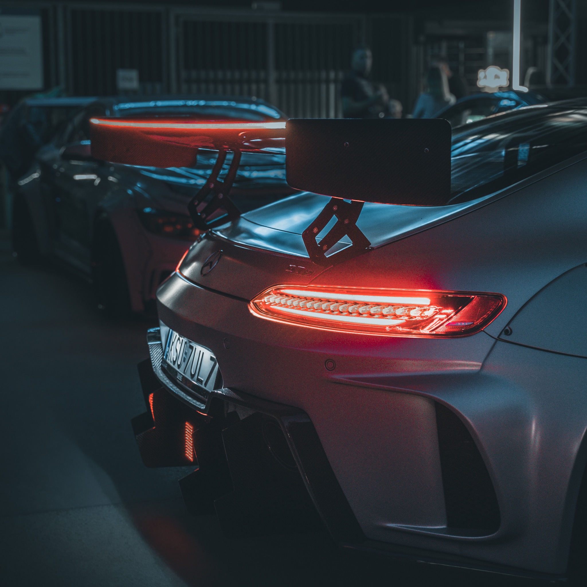 Tył szarego samochodu sportowego z wyraźnie świecącymi tylnymi światłami LED i dużym czarnym spoilerem, tablica rejestracyjna widoczna, w tle inny samochód i osoby.