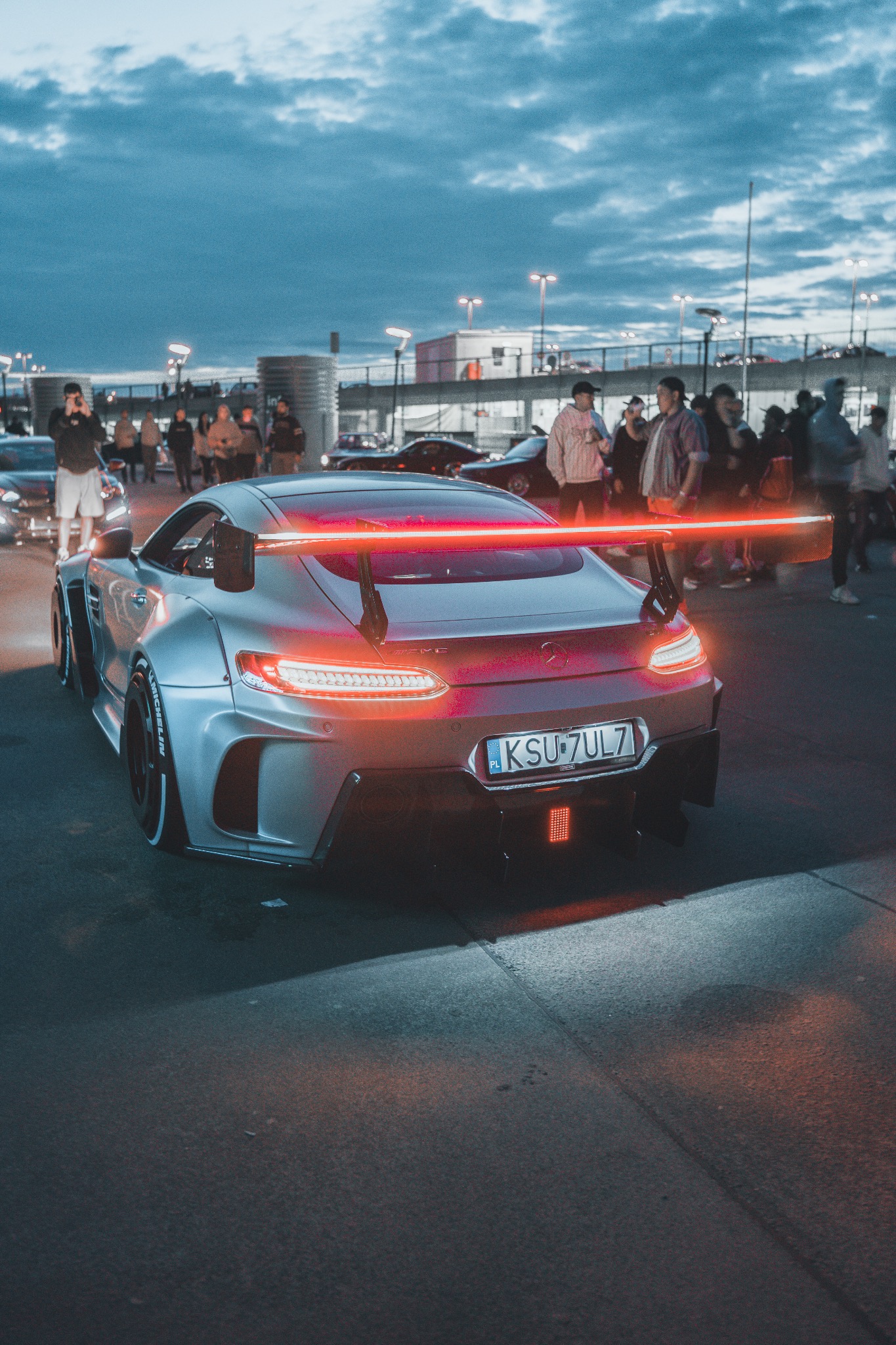 Srebrny Mercedes-AMG GT z neonowym spojlerem i tablicą rejestracyjną KSU 7UL7, sfotografowany w Krakowie na tle grupy obserwujących osób i innych samochodów.