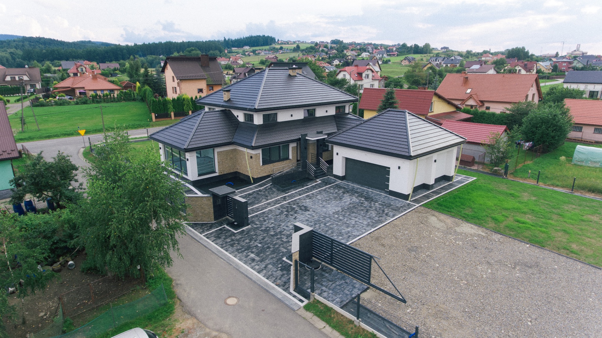 Nowoczesny dom jednorodzinny z ciemną dachówką i jasną elewacją, widok z góry na posesję z kostką brukową i żwirowym podjazdem, w tle zabudowa jednorodzinna i zalesione wzgórza.