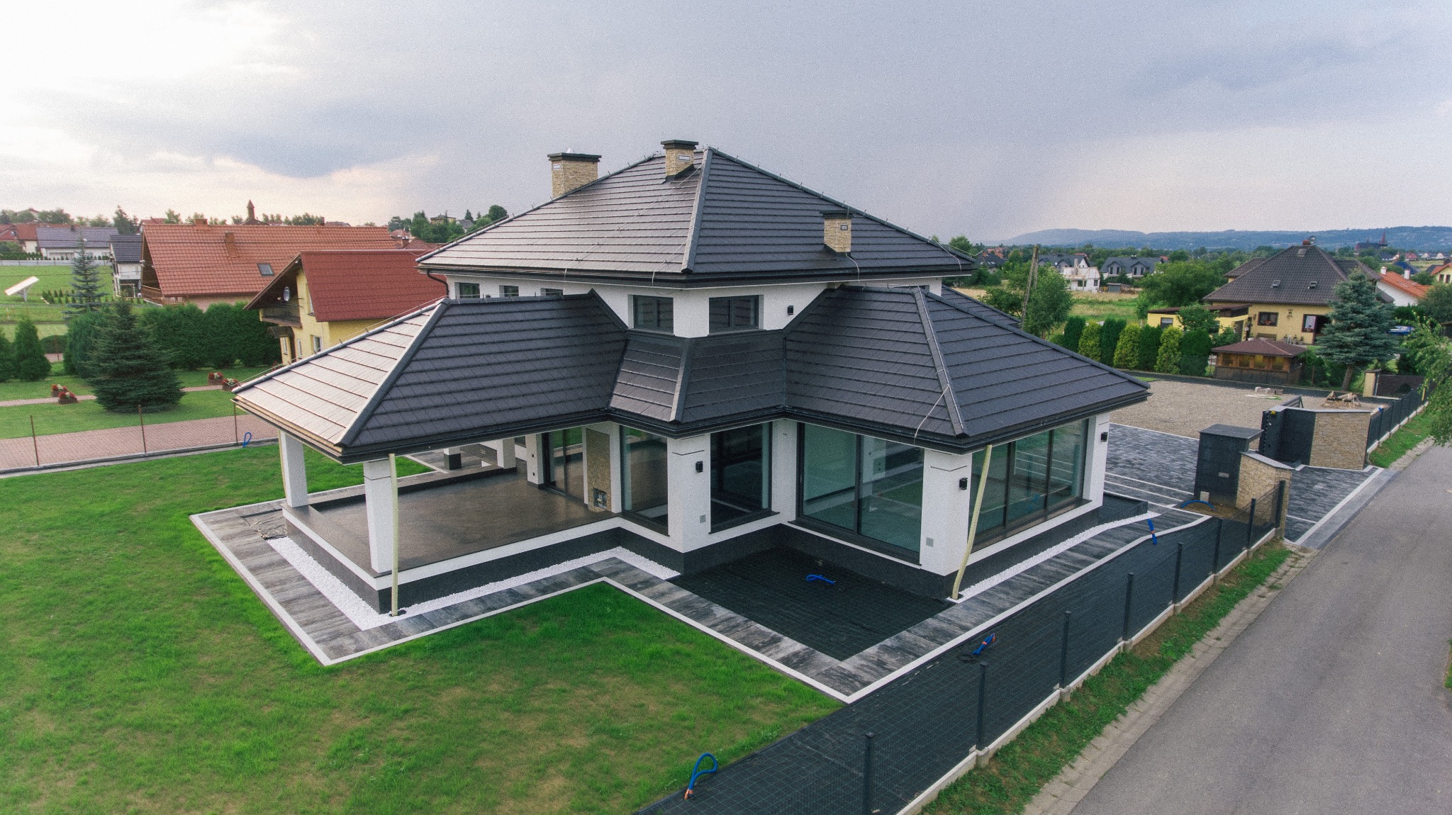Zdjęcie z lotu ptaka nowoczesnego domu jednorodzinnego z ciemną dachówką, dużymi oknami i zadbanym trawnikiem, widoczne inne domy w tle.