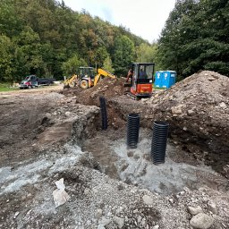 Tartex - Wykonanie wykopu pod instalację sanitarną z widocznymi trzema czarnymi rurami karbowanymi, pomarańczowa mini koparka Hitachi w tle, żółta koparko-ładowarka i samochód dostawczy na skraju lasu.