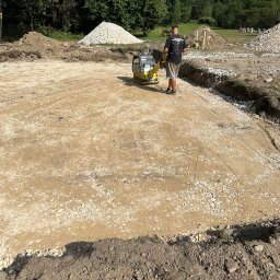 Tartex - Operator zagęszcza podłoże pod budowę za pomocą zagęszczarki wibracyjnej. Widoczne przygotowane wykopy, hałdy ziemi i kruszywa.