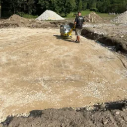 Operator zagęszcza podłoże pod budowę za pomocą zagęszczarki wibracyjnej. Widoczne przygotowane wykopy, hałdy ziemi i kruszywa.