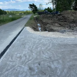 Nowa nawierzchnia z kostki brukowej sąsiadująca z asfaltową drogą, w tle wykop z wodą i sterta ziemi oraz ciężarówka i samochód osobowy