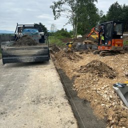 Tartex - Przygotowanie terenu pod budowę; widoczny wykop z wylanym betonem, mini koparka Hitachi, ubijarka, paleta z kostką brukową na ciężarówce, tło z drzewami.