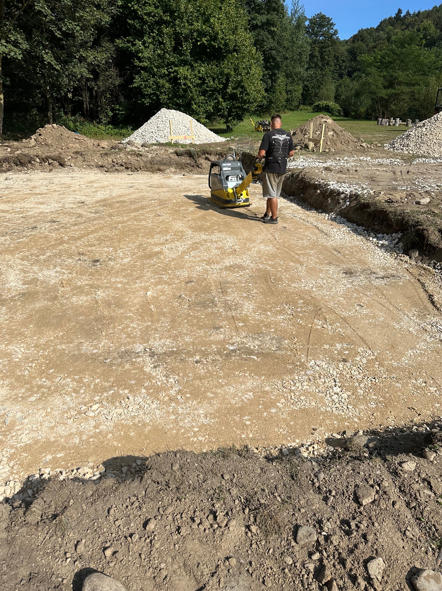Prace ziemne: mężczyzna w szortach zagęszcza podłoże pod budowę zagęszczarką wibracyjną, widoczne pryzmy piasku i kamienia na obrzeżach wykopu.