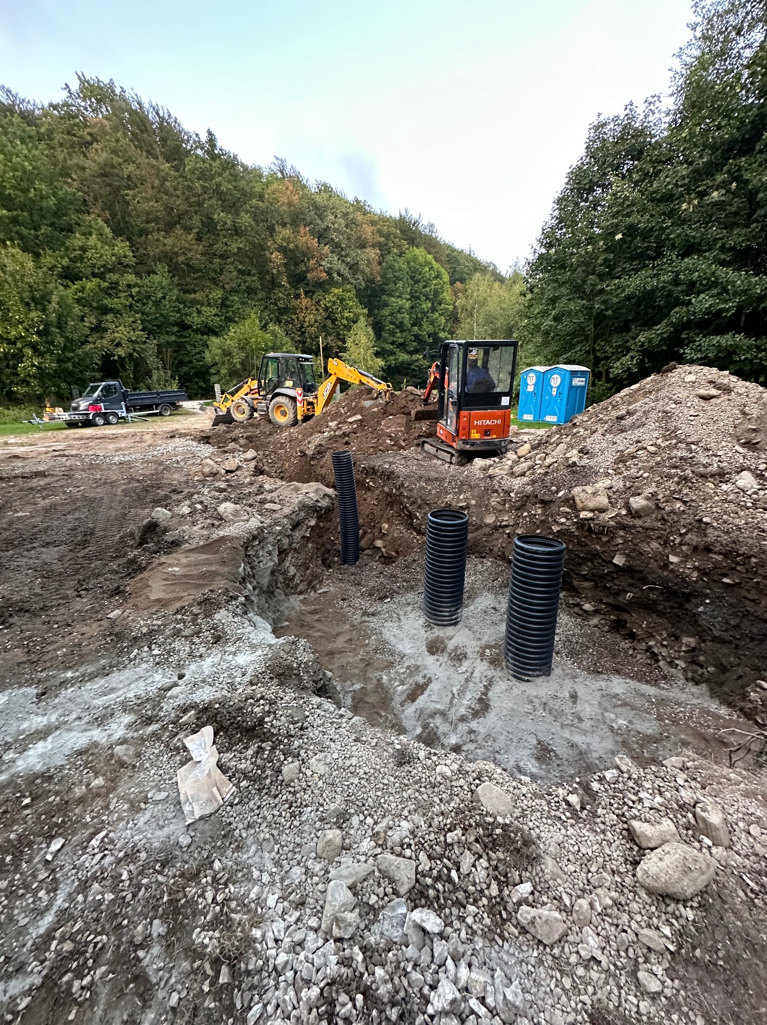 Wykonanie wykopu pod instalację sanitarną z widocznymi trzema czarnymi rurami karbowanymi, pomarańczowa mini koparka Hitachi w tle, żółta koparko-ładowarka i samochód dostawczy na skraju lasu.