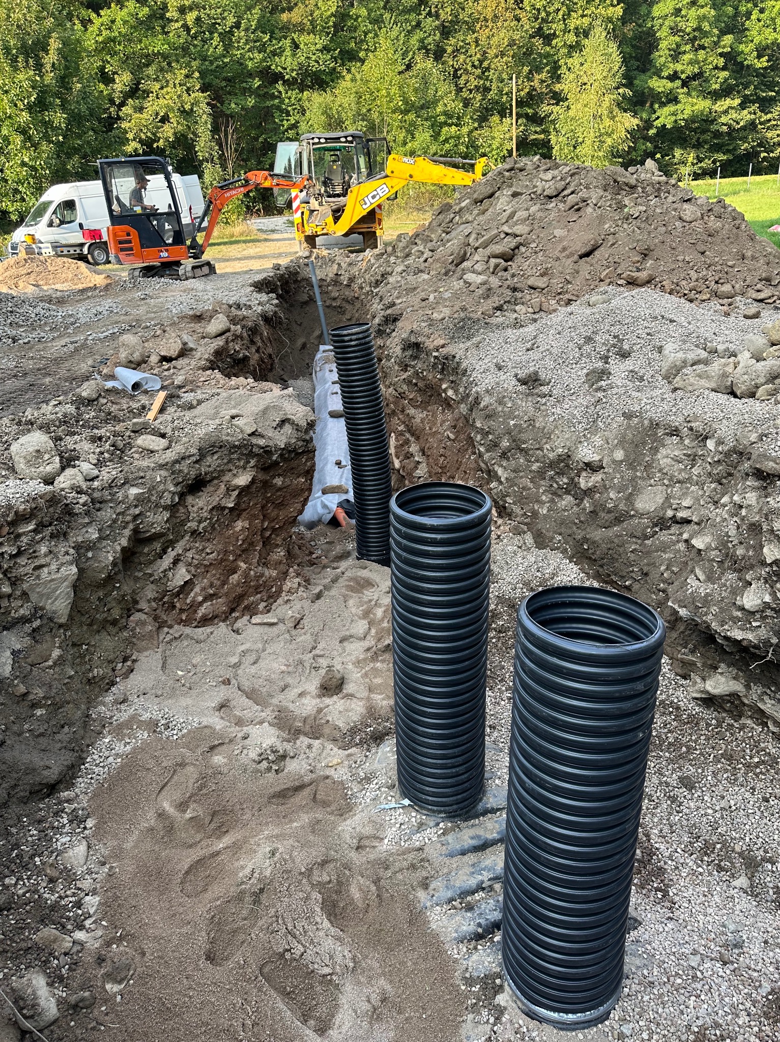 Wykop z ułożonymi rurami drenarskimi, widoczne dwie koparki i furgonetka w tle, ziemia usypana w pryzmę.