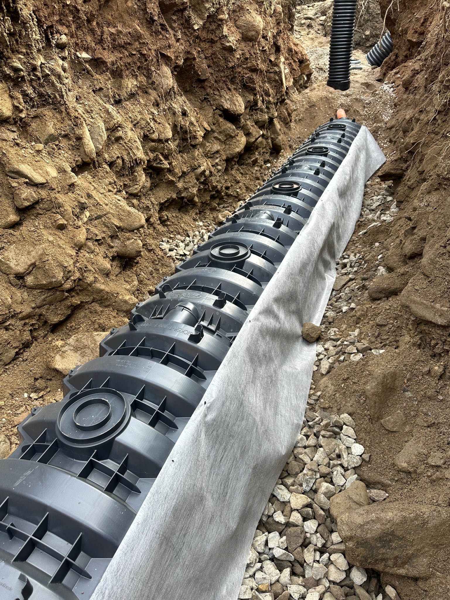 Montaż drenażu rozsączającego z tuneli, otulonego geowłókniną i obsypanego żwirem, w wykopie o gliniastych ścianach.