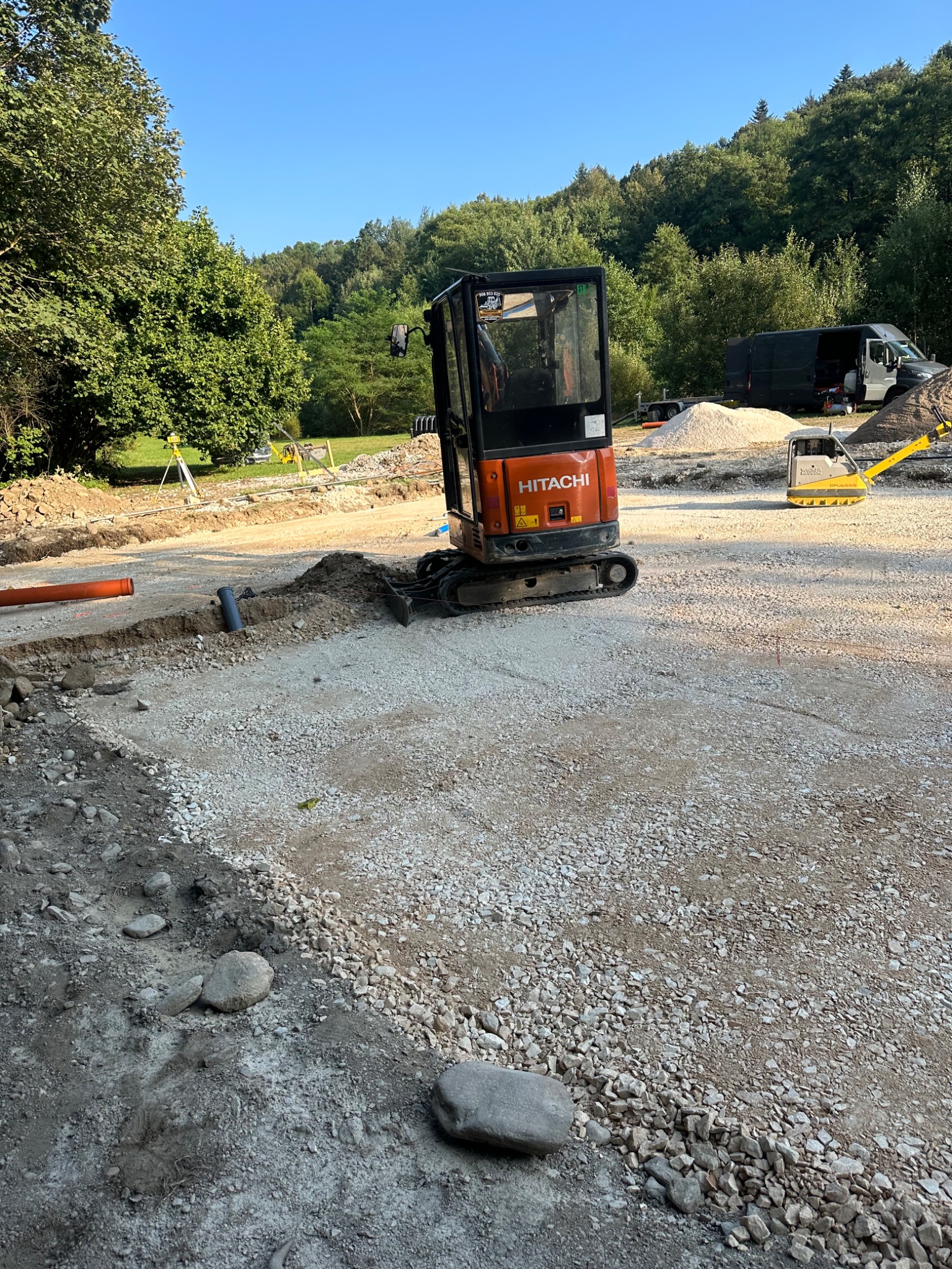 Pomarańczowy minikoparka Hitachi na placu budowy, obok ubijak wibracyjny, widoczne rury i przygotowane podłoże z kruszywa, w tle zieleń drzew i samochód dostawczy.