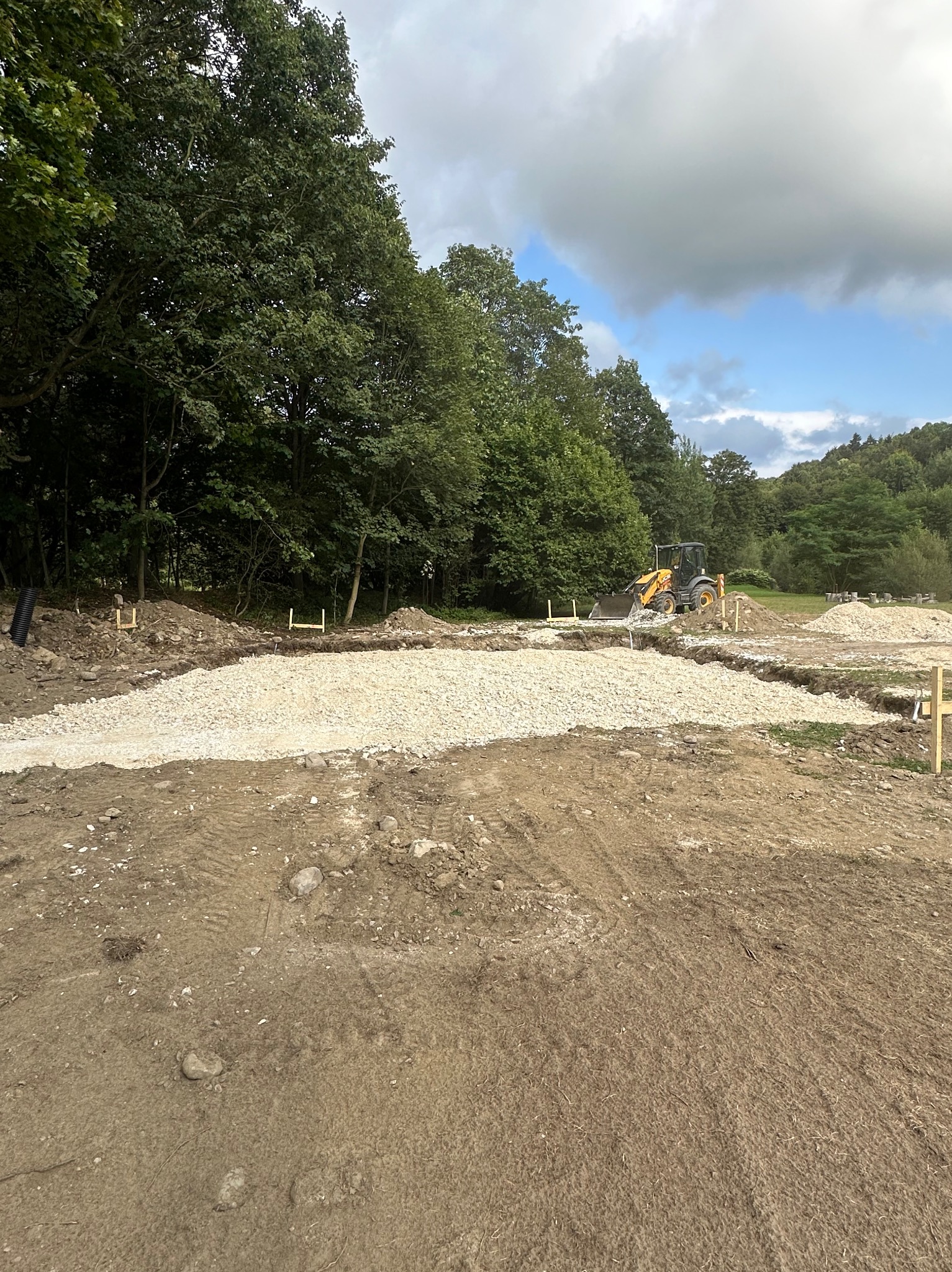 Żółta ładowarka na placu budowy, przygotowanie podłoża z wysypanym żwirem, widoczne drewniane paliki wyznaczające granice, drzewa w tle.