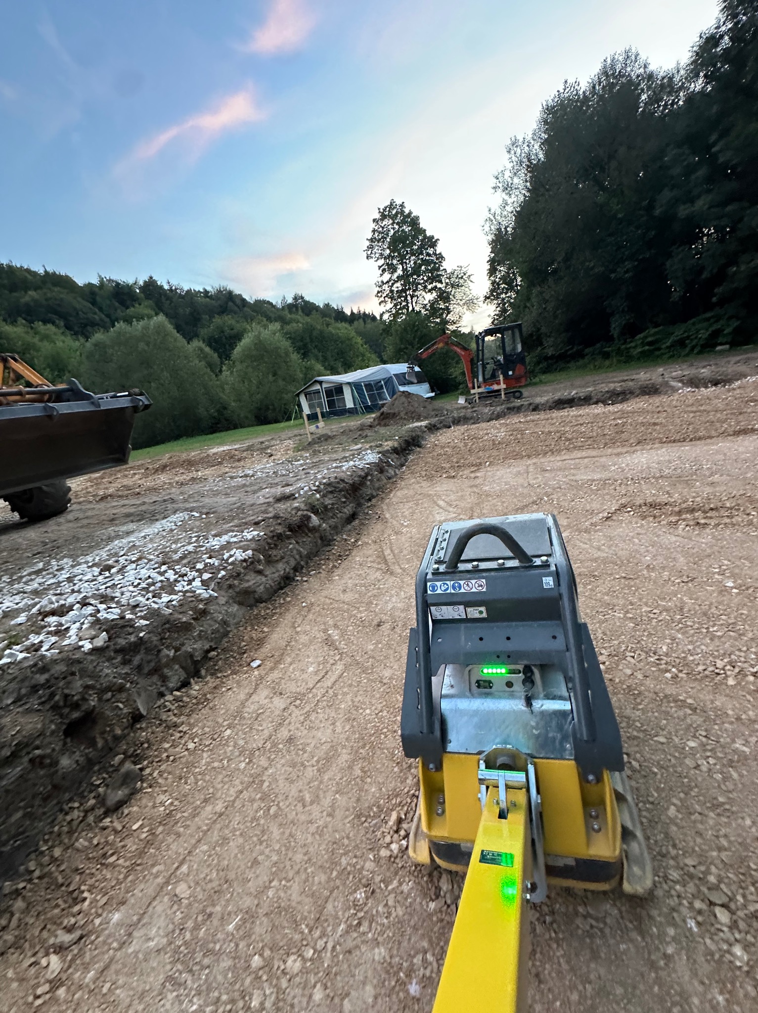 Operator zagęszcza podłoże pod drogę za pomocą ubijarki wibracyjnej; w tle widoczna przyczepa kempingowa, mała koparka i częściowo widoczna koparka z łyżką.