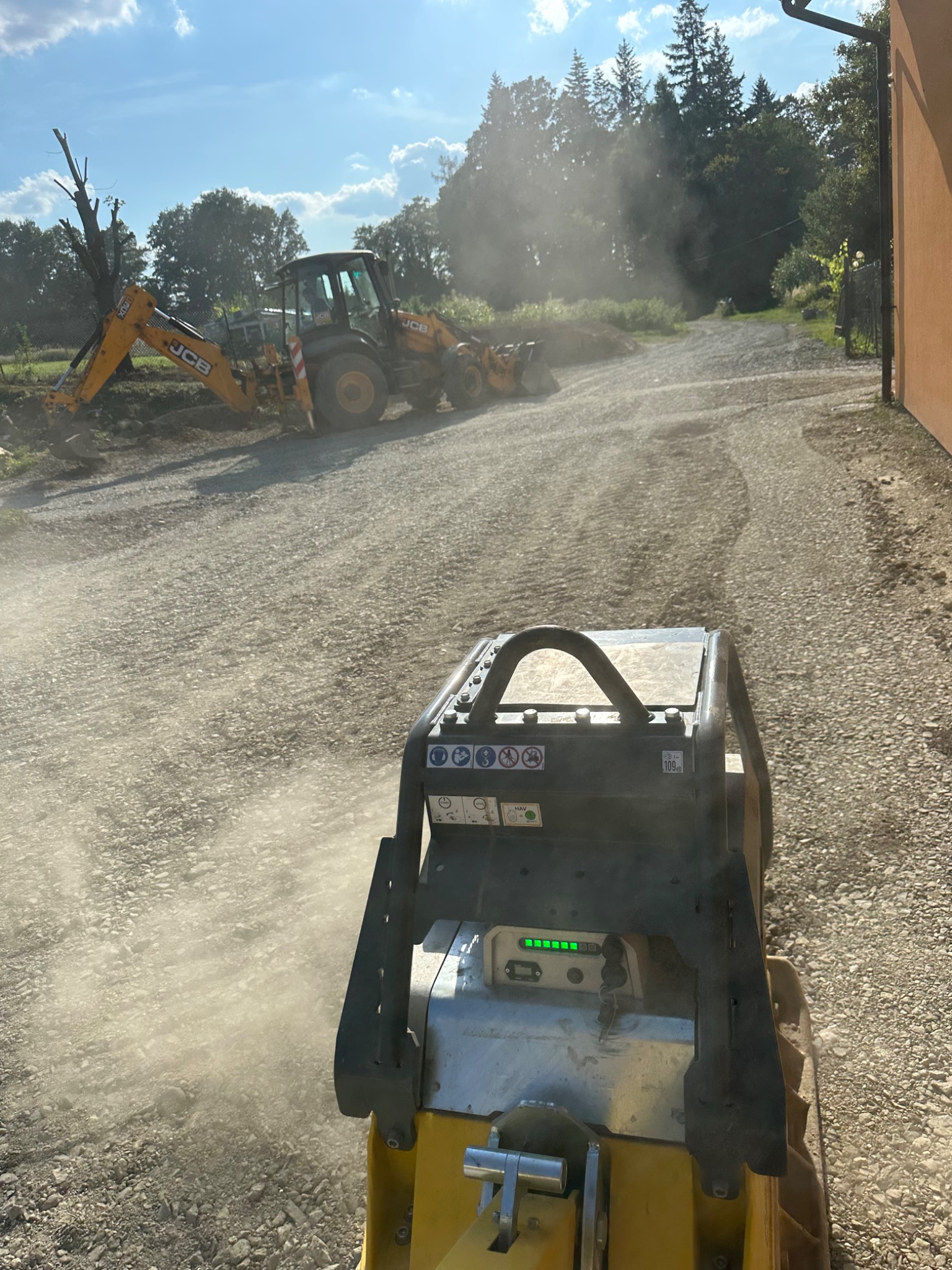 Widok z perspektywy operatora zagęszczarki wibracyjnej na żwirową drogę, w tle koparko-ładowarka JCB i drzewa.