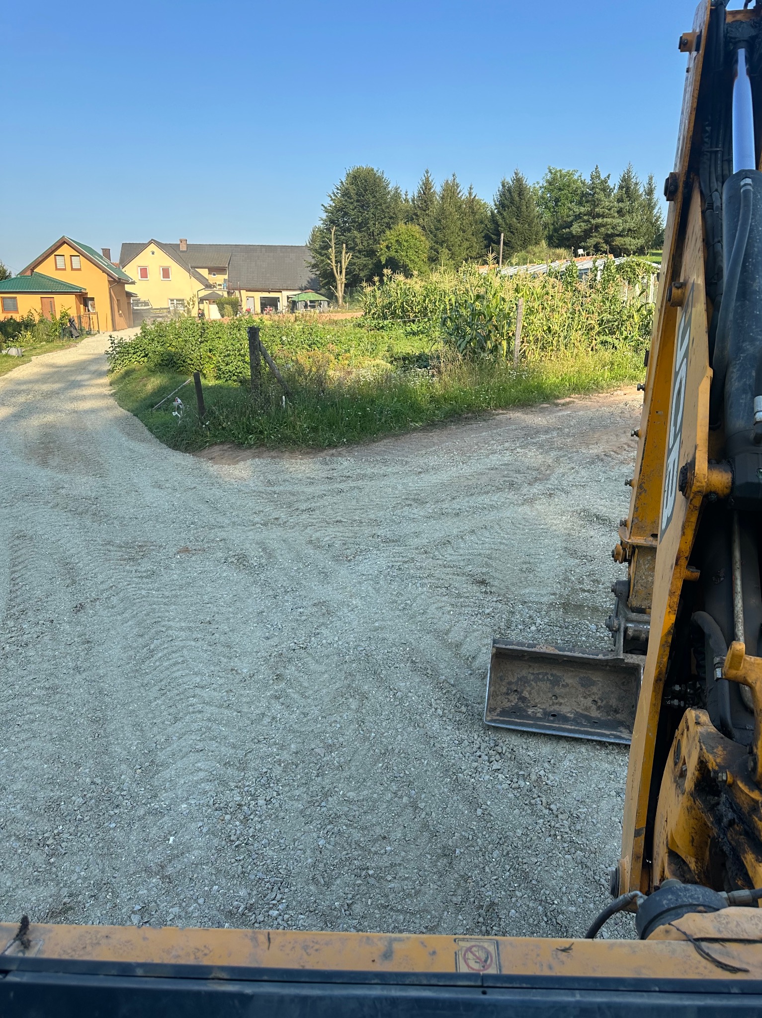 Żwirowa droga rozwidlająca się na tle wiejskich zabudowań, widok z kabiny żółtej maszyny budowlanej z małą łyżką